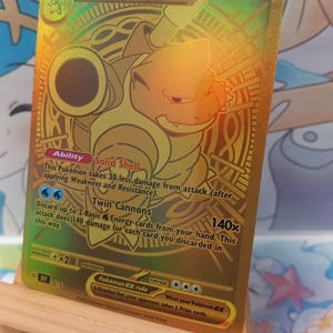 May include: A gold-colored Blastoise ex trading card featuring a metallic finish. The card displays the character Blastoise, 330 HP, the Solid Shell ability, and the Twin Cannons attack. Text includes Pokémon, Stage 2, and game rules.