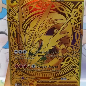 Puede incluir: Una carta coleccionable Pokémon Mega Rayquaza EX de color dorado. La carta presenta una ilustración detallada de una criatura dorada con forma de dragón con el texto "Mega Rayquaza EX" y descripciones de ataque. La carta tiene 320 HP.