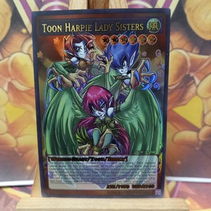 May include: A Toon Harpie Lady Sisters trading card. The card features three cartoonish female figures with wings, in shades of red, blue, and green. The card has a gold border and the text "TOON HARPIE LADY SISTERS" is visible.