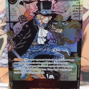 May include: A collectible trading card featuring Sabo from the anime series One Piece. The card showcases Sabo in a black coat and top hat, with a dynamic illustration and text bubbles. The card has a counter value of +1000.