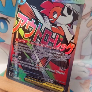 Peut inclure: Une carte à collectionner Mega Scrafty EX avec 330 PV, présentant une illustration de personnage rouge et blanc, le texte japonais アウトローレッグ, ainsi que la capacité Counterattacking Crest et l'attaque Outlaw Leg, posée sur un petit chevalet en bois.