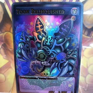 May include: A holographic trading card with the character "Toon Relinquished." The card has a dark blue background with a colourful illustration of a monster. The text "TOON RELINQUISHED" is at the top.