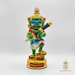 Può includere: Una statua vibrante di un demone tailandese, con un volto verde, dettagli rossi e dorati e un'acconciatura elaborata. La figura tiene in mano un'arma nera e si erge su una base dorata con scritte tailandesi. Rappresentazione della cultura thailandese.