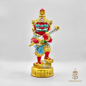 Può includere: Statua vibrante di una figura mitica, prevalentemente rossa con dettagli dorati. La figura ha un copricapo dettagliato, abiti ornati e tiene in mano un'arma color argento. La base è dorata con dettagli incisi. Il volto della figura è verde e bianco.