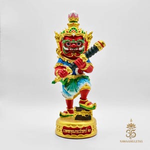 Può includere: Una vivace statuetta rossa e oro di una creatura mitica, che tiene in mano un'arma nera e oro. La figura ha un'espressione feroce, ornata da una corona di gioielli e dettagli intricati. La base è dorata con testo.