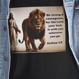 Puede incluir: Camiseta negra con una ilustración de león y el texto "Be strong & courageous, for the Lord, your God, is with you wherever you go. Joshua 1:9". La camiseta se usa con una chaqueta vaquera azul.