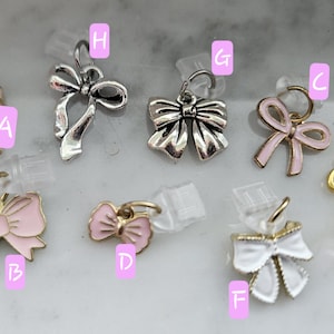 May include: A collection of bow-shaped charms in various colors and finishes. The charms include gold, silver, and pink bows, some with enamel detailing. The bows are attached to small metal loops, suitable for jewelry making or crafting.