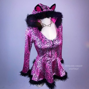 May include: Pink leopard print cat costume with black fur trim, a hood, and a zipper closure. The costume has a heart-shaped neckline and a short skirt.