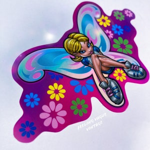 May include: A holographic sticker featuring a cartoon fairy with blonde hair and blue wings sitting on a pink background with colorful flowers. The sticker says "Feeling Vague Vintage" at the bottom.