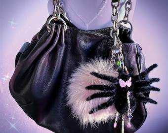 May include: Black leather handbag with silver chain straps. A fluffy pink pom-pom and a black spider charm with a pink bow and dangling beads hang from the bag. The bag has a gathered top and a small pocket on the side.