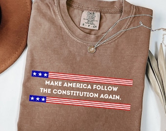 Puede incluir: Una camiseta marrón Comfort Colors con el texto "MAKE AMERICA FOLLOW THE CONSTITUTION AGAIN" en blanco. El texto está encima y debajo de rayas rojas y azules. Un collar plateado con un colgante está sobre la camiseta.