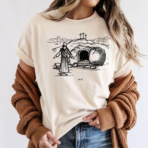 May include: Beige t-shirt featuring a black and white illustration of a religious scene. The graphic depicts a figure with outstretched arms in front of a tomb, with three crosses on a hill in the background. The text "AD 33" is below the illustration.