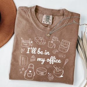 May include: A brown Comfort Colors t-shirt with the text "I'll be in my office" and baking-themed illustrations. A silver necklace with a pendant is draped over the shirt. The shirt has a crew neck and short sleeves.