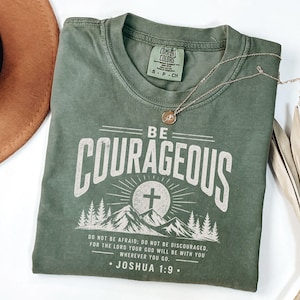 Könnte beinhalten: Olivgrünes T-Shirt mit dem weißen Schriftzug "BE COURAGEOUS". Das Design zeigt ein Kreuz, Berge und den Text "DO NOT BE AFRAID... JOSHUA 1:9." Eine goldene Halskette mit Anhänger liegt auf dem T-Shirt.