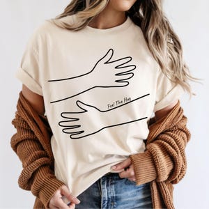 Puede incluir: Camiseta beige con un dibujo lineal negro de dos manos abrazándose. El texto "Feel The Hug" está impreso en negro debajo de las manos. La camiseta está hecha de un material suave.