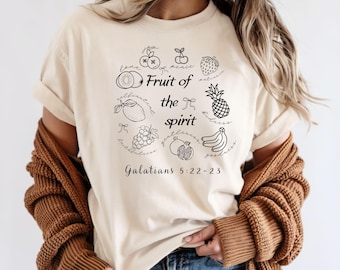 Może przedstawiać: Kremowa koszulka z okrągłym wzorem różnych owoców i napisem "Fruit of the Spirit" czarnym pismem. Tekst "Galatians 5:22-23" jest wydrukowany poniżej. Koszulka ma krótkie rękawy.