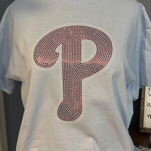 Puede incluir: Camiseta azul claro con un gran logotipo "P" adornado con pedrería. El logotipo está delineado en blanco y relleno con pedrería rosa y roja. La camiseta se muestra en un maniquí.