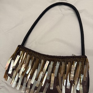 Puede incluir: Un bolso de mano de satén marrón con un asa negra. La parte superior del bolso está adornada con pequeñas cuentas doradas y detalles rectangulares de nácar que cuelgan. El bolso tiene un cierre de cremallera.