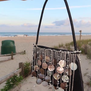 Peut inclure: Un sac à main noir doté d'une anse unique et d'un panneau avant décoratif orné de brins verticaux de perles transparentes entremêlés de disques circulaires iridescents en nacre qui pendent librement du bord supérieur du sac.