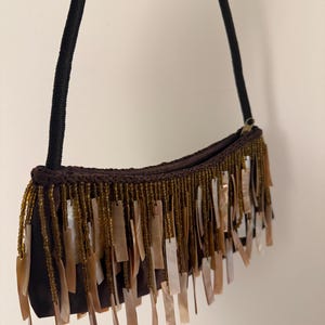 May include: A brown suede shoulder bag with a black rope strap. The bag is decorated with gold beads and rectangular shell accents, creating a fringed effect. The bag has a zip closure.