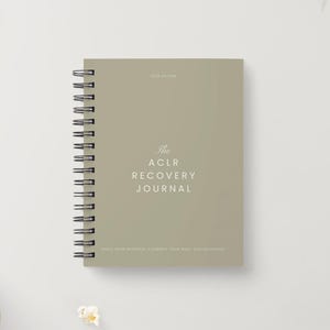 Peut inclure: Un journal à spirales vert sauge avec le texte "The ACLR Recovery Journal" en écriture blanche. Le journal est posé sur une surface blanche, avec un crayon et un bol de pop-corn en arrière-plan.