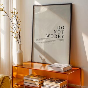 May include: A living room scene with a framed print displaying the words "DO NOT WORRY." An orange cabinet holds a vase with yellow-flowered branches and several books. A yellow armchair is visible in the foreground, with sunlight streaming through the window.