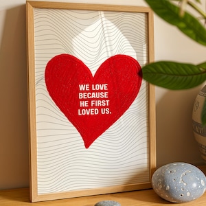 Op de afbeelding: Een ingelijste kunstafdruk met een groot rood hart en de tekst "WE LOVE BECAUSE HE FIRST LOVED US." Het hart staat op een achtergrond van golvende grijze lijnen. De lijst is van lichtkleurig hout.