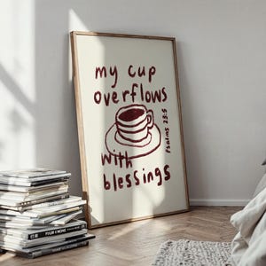 May include: A framed art print with the words "my cup overflows with blessings" in burgundy script. The artwork features a burgundy coffee cup illustration. The print is displayed on a wooden floor, leaning against a white wall.