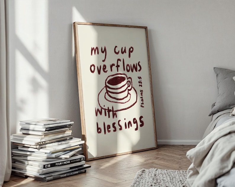 May include: A framed art print with the words "my cup overflows with blessings" in burgundy script. The artwork features a burgundy coffee cup illustration. The print is displayed on a wooden floor, leaning against a white wall.