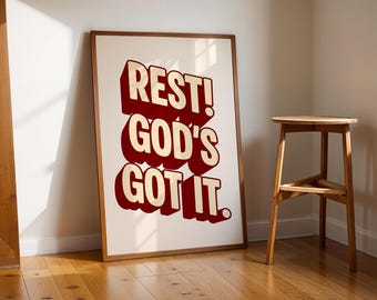 May include: A framed print with the words "REST! GOD'S GOT IT." in a bold, 3D-style font. The text is red with a cream-colored outline, set against a white background. The frame is a warm brown. A wooden stool is to the right.