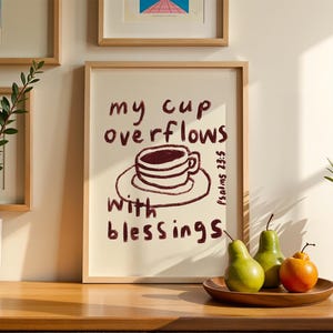 Könnte beinhalten: Ein gerahmter Druck mit den Worten "my cup overflows with blessings" und einer Zeichnung einer Kaffeetasse. Der Druck befindet sich in einem hellen Holzrahmen. Zwei grüne Birnen und eine orangefarbene Frucht liegen auf einem Holztablett.