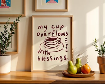 May include: A framed print with the words "my cup overflows with blessings" and a drawing of a coffee cup. The print is in a light wood frame. Two green pears and an orange fruit sit on a wooden tray.