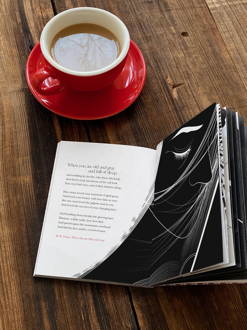 May include: An open book with a black and white illustration of a woman's face on the right page. The left page features a poem by W.B. Yeats titled "When You Are Old and Gray". The book is lying on a wooden table with a red coffee cup and saucer.