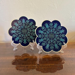May include: Two navy blue ceramic coasters with a floral design in teal and white. The coasters are shaped like a flower with a scalloped edge. They are displayed on clear plastic stands on a wooden surface.