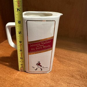 May include: White ceramic pitcher with a handle and a spout. The front features a rectangular label with "Johnnie Walker Red Label Old Scotch Whisky" text and a walking figure. The pitcher is approximately 7 inches tall.