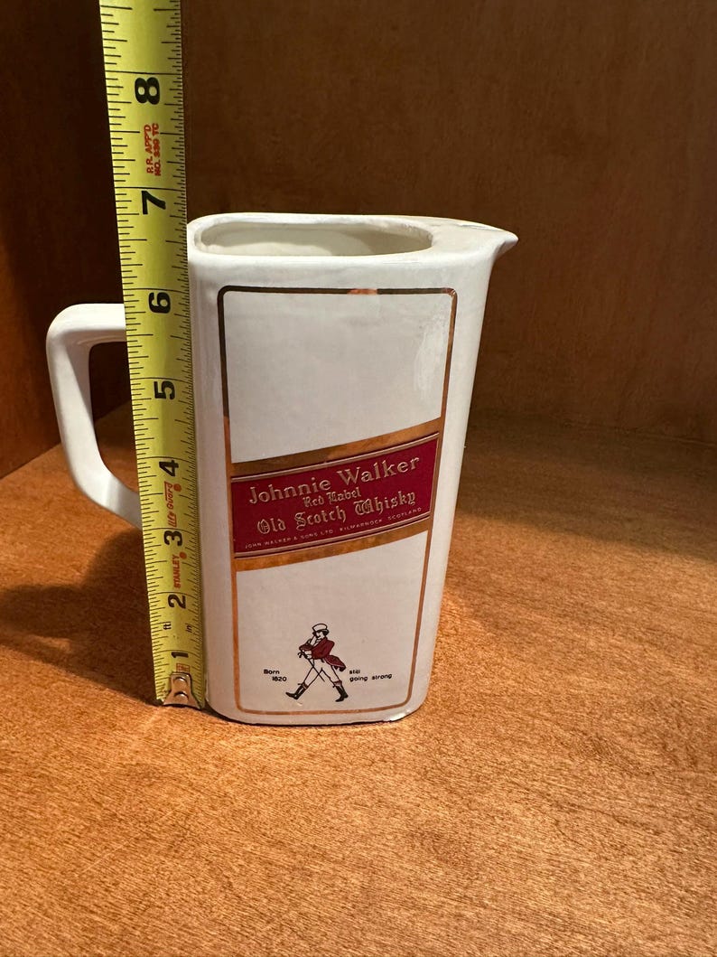 May include: White ceramic pitcher with a handle and a spout. The front features a rectangular label with "Johnnie Walker Red Label Old Scotch Whisky" text and a walking figure. The pitcher is approximately 7 inches tall.