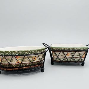 May include: Two ceramic serving dishes with floral patterns, set in black metal wire holders. One is oval, the other rectangular. The dishes have a cream-colored interior and a green and brown floral design.