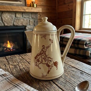 May include: Cream-colored ceramic coffee pot with a brown cowboy and horse design. The pot has a lid, a curved handle, and a spout. The design features a cowboy riding a horse, with rope and horseshoe details.
