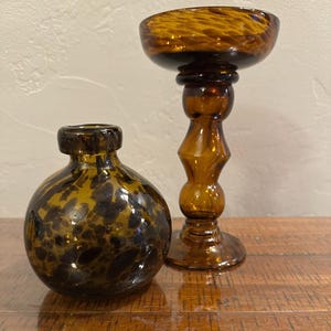 May include: A small, round, amber-colored glass vase with a dark brown mottled pattern and a narrow neck, positioned next to a tall, amber-colored glass candle holder featuring a wide, bowl-shaped top and a decorative, multi-tiered pedestal base.