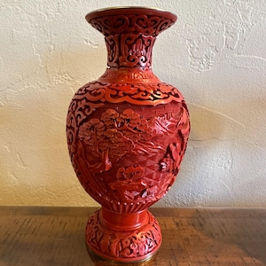May include: A cinnabar red vase featuring intricate relief carvings of a landscape with trees, mountains, and architectural elements. The vase has a flared rim, a bulbous body, and a circular base, all finished with a metallic trim at the top and bottom.
