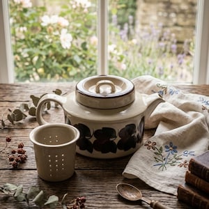 May include: Cream-colored ceramic teapot with a matching infuser and lid, adorned with dark blue floral patterns. A vintage spoon and embroidered linen napkin with blue flowers sit on a rustic wooden table, creating a cozy, farmhouse aesthetic.