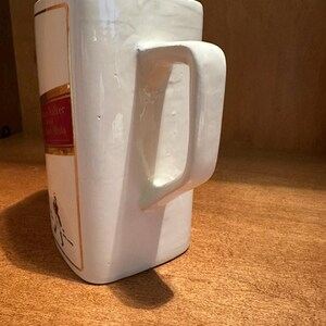 May include: A white ceramic mug with a rectangular shape and a large handle. The mug features a red and gold label with text and a figure. The mug is sitting on a wooden surface.