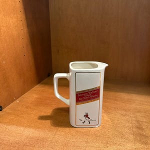 May include: White ceramic pitcher with a rectangular shape and a handle. The pitcher features the "Johnnie Walker Old Scotch Whisky" logo in red and gold, with a figure of a striding man. A classic barware item.