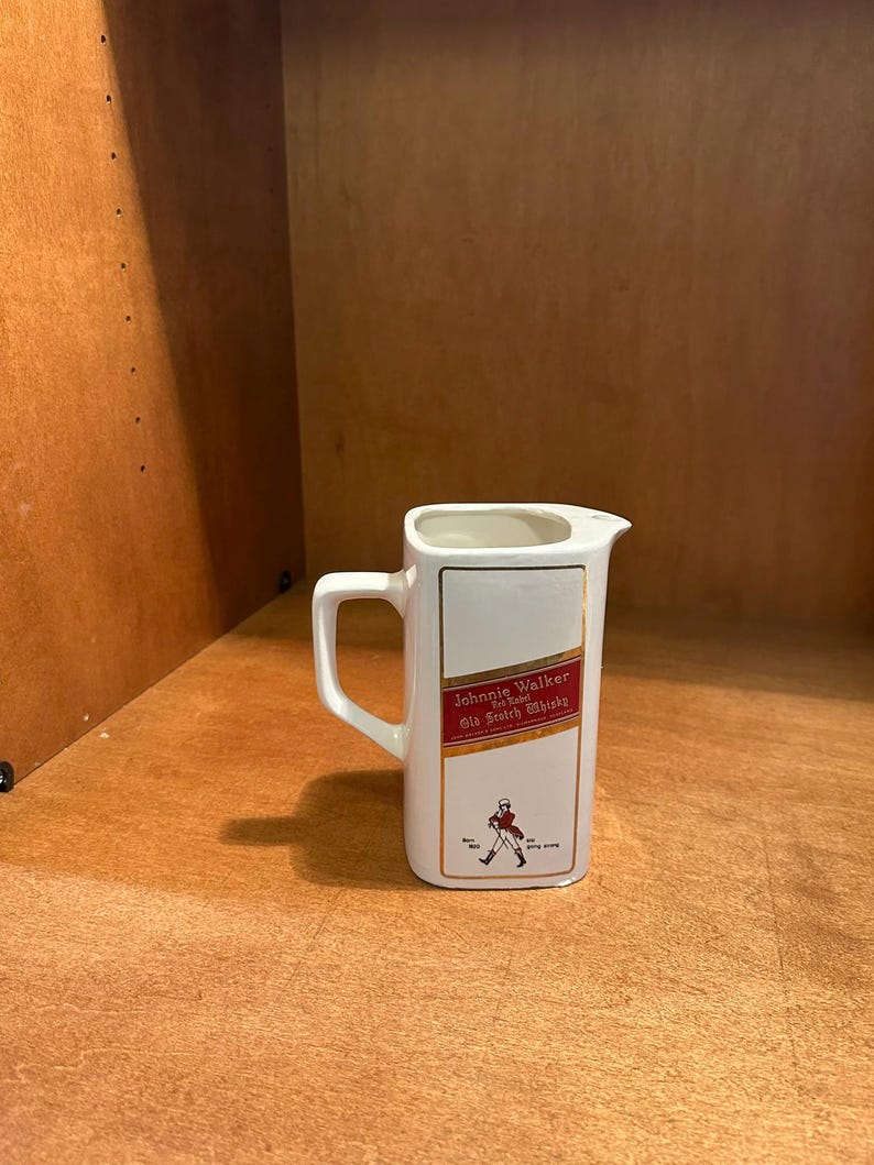 May include: White ceramic pitcher with a rectangular shape and a handle. The pitcher features the "Johnnie Walker Old Scotch Whisky" logo in red and gold, with a figure of a striding man. A classic barware item.