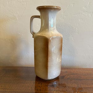 May include: A ceramic pitcher with a hexagonal body and a single handle. The vessel features a gradient glaze that transitions from a deep, warm brown at the base to a lighter, creamy beige tone toward the flared rim and the handle attachment point.