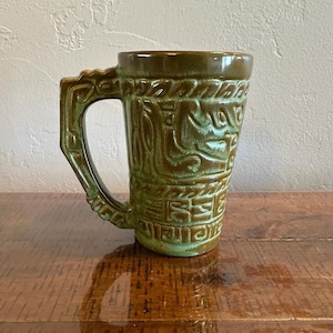May include: A ceramic mug featuring a deep olive green glaze with lighter green highlights. The exterior surface is decorated with a raised, textured relief pattern of geometric shapes and abstract lines. It has a sturdy, angular handle.