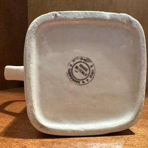 May include: A white ceramic pitcher with a square base and a small spout. The bottom of the pitcher has a circular stamp that reads "MADE IN ITALY". The pitcher is sitting on a wooden surface.