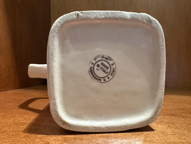 May include: A white ceramic pitcher with a square base and a small spout. The bottom of the pitcher has a circular stamp that reads "MADE IN ITALY". The pitcher is sitting on a wooden surface.