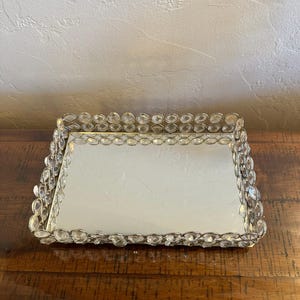 May include: A rectangular vanity tray featuring a reflective mirrored base and a raised border composed of clear, faceted, oval-shaped glass crystals set in a silver-toned metal frame. The tray has a flat surface and a decorative, textured edge.