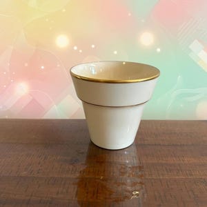 May include: A cream-colored ceramic cup with a flared rim and a thin gold band along the top edge. The cup features a smooth, solid finish and a tapered body that narrows toward the base, presenting a simple, functional design for holding liquids.